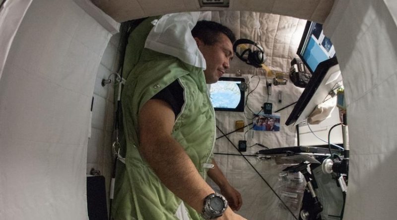 An astronaut inside a spacecraft, standing upright in a green sleeping bag-like garment. The compact interior features equipment and controls on the walls and ceiling, with a small window in front letting in light. The image has a fisheye lens effect, creating a slightly distorted perspective.