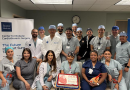 A group of medical professionals, including doctors and nurses, pose in a hospital setting to celebrate a milestone. They stand around a cake that reads '800 Robotic Heart'. Behind them are banners for 'Baylor Medicine' and the 'Center for Robotic Cardiothoracic Surgery'. The room features fluorescent lighting and a wall clock.