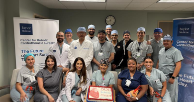 A group of medical professionals, including doctors and nurses, pose in a hospital setting to celebrate a milestone. They stand around a cake that reads '800 Robotic Heart'. Behind them are banners for 'Baylor Medicine' and the 'Center for Robotic Cardiothoracic Surgery'. The room features fluorescent lighting and a wall clock.