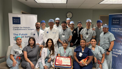 A group of medical professionals, including doctors and nurses, pose in a hospital setting to celebrate a milestone. They stand around a cake that reads '800 Robotic Heart'. Behind them are banners for 'Baylor Medicine' and the 'Center for Robotic Cardiothoracic Surgery'. The room features fluorescent lighting and a wall clock.