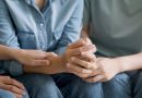 A close-up of several people sitting side by side in casual clothing, such as denim jeans and shirts. Their hands are gently intertwined, conveying a sense of comfort, support, and emotional connection. The image emphasizes themes of empathy, unity, and human solidarity.
