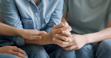 A close-up of several people sitting side by side in casual clothing, such as denim jeans and shirts. Their hands are gently intertwined, conveying a sense of comfort, support, and emotional connection. The image emphasizes themes of empathy, unity, and human solidarity.
