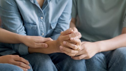 A close-up of several people sitting side by side in casual clothing, such as denim jeans and shirts. Their hands are gently intertwined, conveying a sense of comfort, support, and emotional connection. The image emphasizes themes of empathy, unity, and human solidarity.