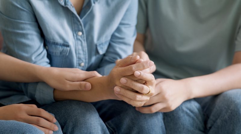 A close-up of several people sitting side by side in casual clothing, such as denim jeans and shirts. Their hands are gently intertwined, conveying a sense of comfort, support, and emotional connection. The image emphasizes themes of empathy, unity, and human solidarity.