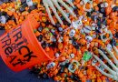 An orange plastic bucket labeled 'TRICK OR TREAT' is tipped over, spilling assorted Halloween candies including candy corn, wrapped chocolates, lollipops, and sweets in orange and black wrappers. Small plastic skulls and skeleton hands are mixed in, creating a festive Halloween scene.