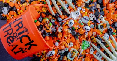 An orange plastic bucket labeled 'TRICK OR TREAT' is tipped over, spilling assorted Halloween candies including candy corn, wrapped chocolates, lollipops, and sweets in orange and black wrappers. Small plastic skulls and skeleton hands are mixed in, creating a festive Halloween scene.