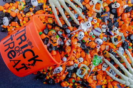 An orange plastic bucket labeled 'TRICK OR TREAT' is tipped over, spilling assorted Halloween candies including candy corn, wrapped chocolates, lollipops, and sweets in orange and black wrappers. Small plastic skulls and skeleton hands are mixed in, creating a festive Halloween scene.