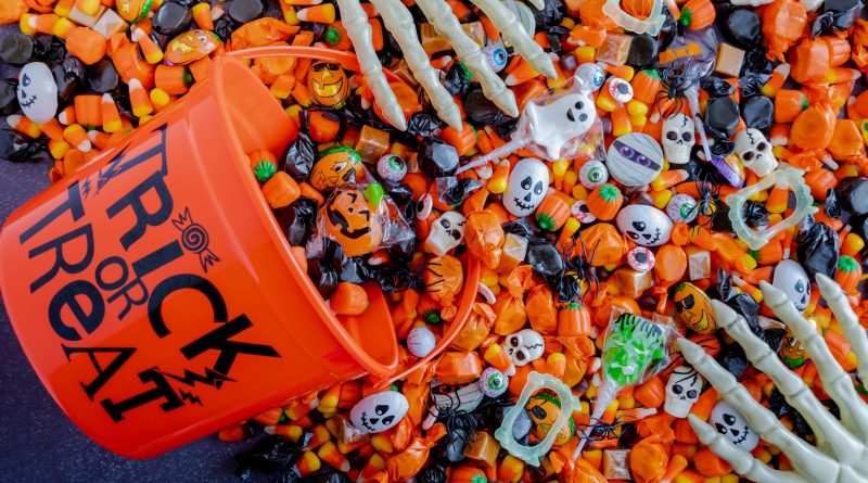 An orange plastic bucket labeled 'TRICK OR TREAT' is tipped over, spilling assorted Halloween candies including candy corn, wrapped chocolates, lollipops, and sweets in orange and black wrappers. Small plastic skulls and skeleton hands are mixed in, creating a festive Halloween scene.