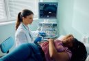 A healthcare professional in a white coat and blue gloves performs an abdominal ultrasound on a patient lying on an examination table. The patient rests one arm on their head and the other by their side. An ultrasound machine displays imaging on a monitor nearby, with natural light entering through a window with partially open blinds.