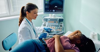 A healthcare professional in a white coat and blue gloves performs an abdominal ultrasound on a patient lying on an examination table. The patient rests one arm on their head and the other by their side. An ultrasound machine displays imaging on a monitor nearby, with natural light entering through a window with partially open blinds.