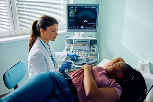 A healthcare professional in a white coat and blue gloves performs an abdominal ultrasound on a patient lying on an examination table. The patient rests one arm on their head and the other by their side. An ultrasound machine displays imaging on a monitor nearby, with natural light entering through a window with partially open blinds.
