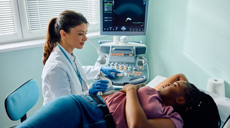 A healthcare professional in a white coat and blue gloves performs an abdominal ultrasound on a patient lying on an examination table. The patient rests one arm on their head and the other by their side. An ultrasound machine displays imaging on a monitor nearby, with natural light entering through a window with partially open blinds.