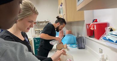 A group of DNP students practice airway management techniques in a clinical simulation setting. One individual uses a bag-valve mask to ventilate a medical mannequin, while another works on a separate mannequin, possibly performing intubation. The room is equipped with medical supplies, red biohazard containers, and organized storage cabinets, emphasizing a hands-on learning environment for emergency care procedures.