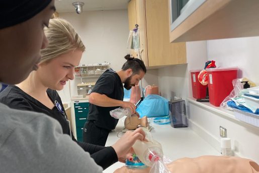 A group of DNP students practice airway management techniques in a clinical simulation setting. One individual uses a bag-valve mask to ventilate a medical mannequin, while another works on a separate mannequin, possibly performing intubation. The room is equipped with medical supplies, red biohazard containers, and organized storage cabinets, emphasizing a hands-on learning environment for emergency care procedures.