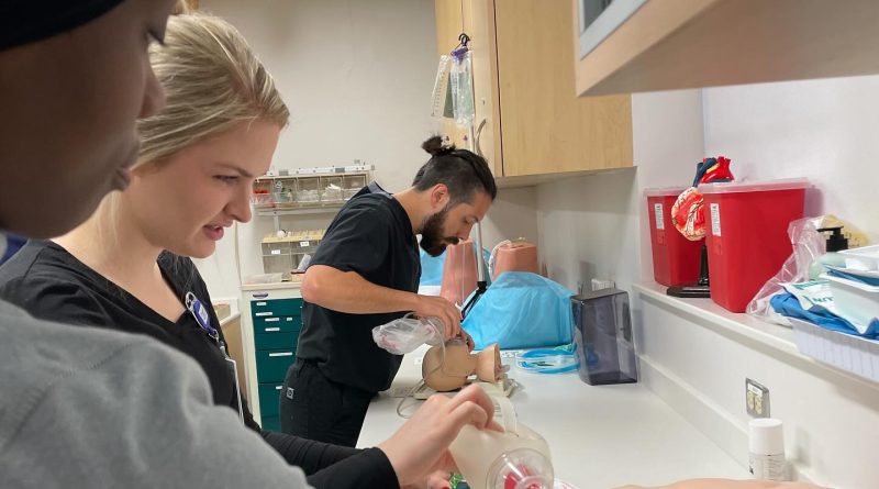 A group of DNP students practice airway management techniques in a clinical simulation setting. One individual uses a bag-valve mask to ventilate a medical mannequin, while another works on a separate mannequin, possibly performing intubation. The room is equipped with medical supplies, red biohazard containers, and organized storage cabinets, emphasizing a hands-on learning environment for emergency care procedures.
