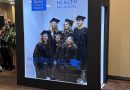 A large display box featuring a group of graduates wearing black caps and gowns with gold accents. The backdrop inside the box shows the Baylor College of Medicine logo and the text “School of Health Professions.” Additional text reads “Congratulations, Class of 2025” alongside a blue graduation cap graphic. The display is set in an indoor hallway with beige walls and patterned carpet, and there are holiday garlands visible in the background.