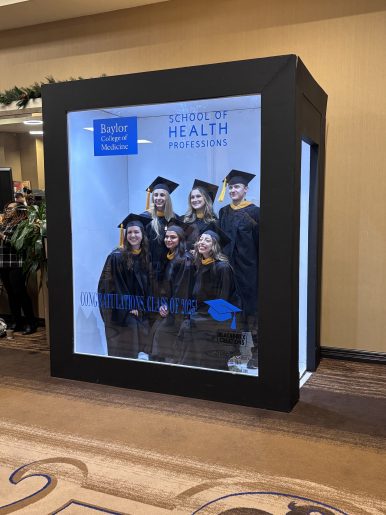 A large display box featuring a group of graduates wearing black caps and gowns with gold accents. The backdrop inside the box shows the Baylor College of Medicine logo and the text “School of Health Professions.” Additional text reads “Congratulations, Class of 2025” alongside a blue graduation cap graphic. The display is set in an indoor hallway with beige walls and patterned carpet, and there are holiday garlands visible in the background.