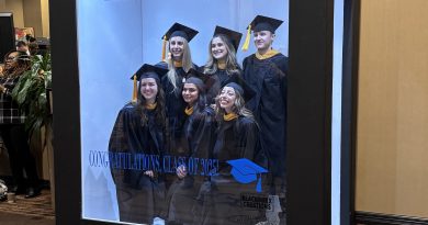 A large display box featuring a group of graduates wearing black caps and gowns with gold accents. The backdrop inside the box shows the Baylor College of Medicine logo and the text “School of Health Professions.” Additional text reads “Congratulations, Class of 2025” alongside a blue graduation cap graphic. The display is set in an indoor hallway with beige walls and patterned carpet, and there are holiday garlands visible in the background.