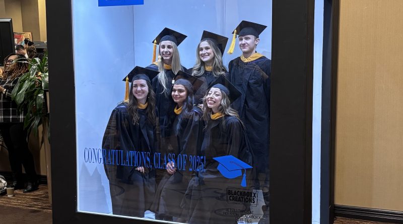 A large display box featuring a group of graduates wearing black caps and gowns with gold accents. The backdrop inside the box shows the Baylor College of Medicine logo and the text “School of Health Professions.” Additional text reads “Congratulations, Class of 2025” alongside a blue graduation cap graphic. The display is set in an indoor hallway with beige walls and patterned carpet, and there are holiday garlands visible in the background.