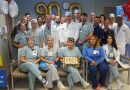 A group of medical professionals wearing scrubs and white coats gathered in a hospital or clinic setting for a celebration. They are posing around a rectangular cake with a decorative design placed on a chair in the front row. Behind them, gold balloon numbers “900” are displayed on the wall, along with a clock above. Red and white star-shaped balloons are positioned on the right side, and a Baylor Medicine banner is partially visible in the background.
