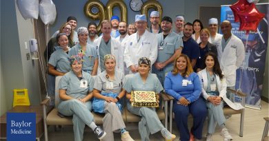 A group of medical professionals wearing scrubs and white coats gathered in a hospital or clinic setting for a celebration. They are posing around a rectangular cake with a decorative design placed on a chair in the front row. Behind them, gold balloon numbers “900” are displayed on the wall, along with a clock above. Red and white star-shaped balloons are positioned on the right side, and a Baylor Medicine banner is partially visible in the background.