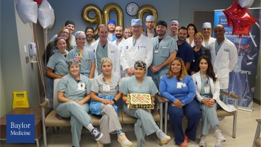 A group of medical professionals wearing scrubs and white coats gathered in a hospital or clinic setting for a celebration. They are posing around a rectangular cake with a decorative design placed on a chair in the front row. Behind them, gold balloon numbers “900” are displayed on the wall, along with a clock above. Red and white star-shaped balloons are positioned on the right side, and a Baylor Medicine banner is partially visible in the background.