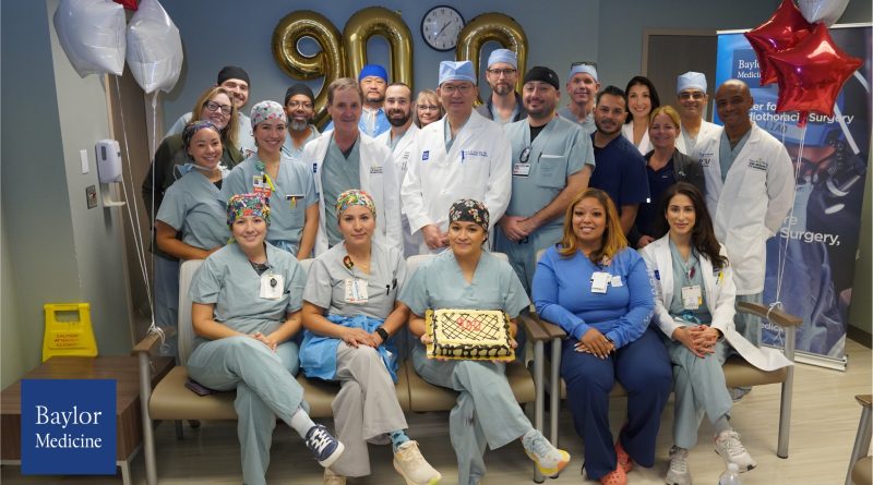 A group of medical professionals wearing scrubs and white coats gathered in a hospital or clinic setting for a celebration. They are posing around a rectangular cake with a decorative design placed on a chair in the front row. Behind them, gold balloon numbers “900” are displayed on the wall, along with a clock above. Red and white star-shaped balloons are positioned on the right side, and a Baylor Medicine banner is partially visible in the background.