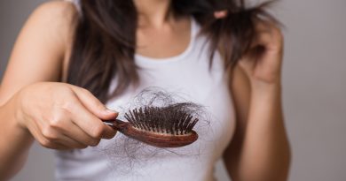 A person holding a wooden hairbrush filled with a large amount of shed hair while using the other hand to examine strands of their own long, dark hair. The individual is wearing a white sleeveless top, and the focus is on the hairbrush and hair loss.