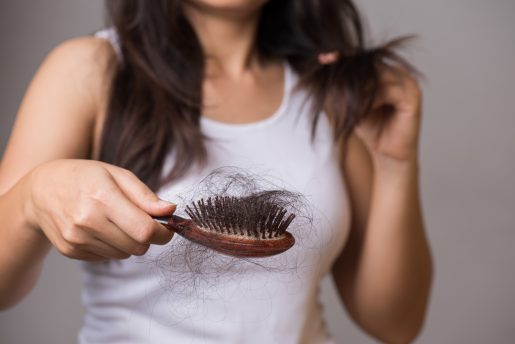 A person holding a wooden hairbrush filled with a large amount of shed hair while using the other hand to examine strands of their own long, dark hair. The individual is wearing a white sleeveless top, and the focus is on the hairbrush and hair loss.