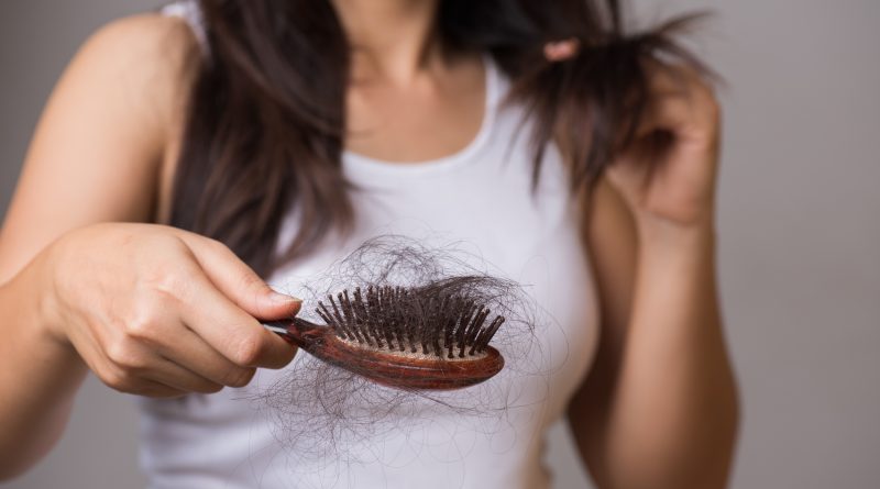 A person holding a wooden hairbrush filled with a large amount of shed hair while using the other hand to examine strands of their own long, dark hair. The individual is wearing a white sleeveless top, and the focus is on the hairbrush and hair loss.