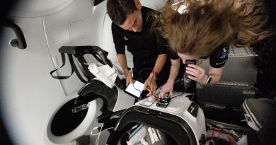 Inside a spacecraft cabin, two individuals float in microgravity while interacting with electronic devices secured to a workstation. One person is leaning over a control surface, using a touchscreen device, while another reviews information on a tablet. Their hair and equipment are suspended due to weightlessness, and the surrounding environment features sleek white panels, seats, and harnesses typical of a crewed space capsule.
