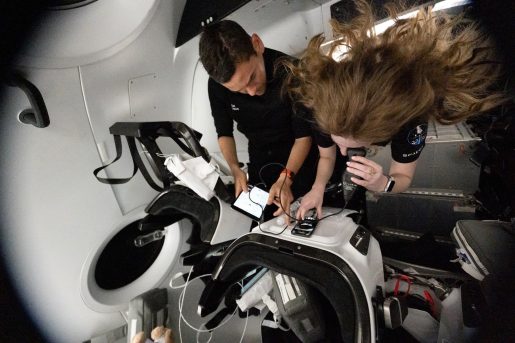 Inside a spacecraft cabin, two individuals float in microgravity while interacting with electronic devices secured to a workstation. One person is leaning over a control surface, using a touchscreen device, while another reviews information on a tablet. Their hair and equipment are suspended due to weightlessness, and the surrounding environment features sleek white panels, seats, and harnesses typical of a crewed space capsule.