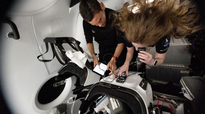 Inside a spacecraft cabin, two individuals float in microgravity while interacting with electronic devices secured to a workstation. One person is leaning over a control surface, using a touchscreen device, while another reviews information on a tablet. Their hair and equipment are suspended due to weightlessness, and the surrounding environment features sleek white panels, seats, and harnesses typical of a crewed space capsule.