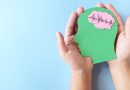 Two hands hold a green paper cutout shaped like a human head against a light blue background, with a pink brain shape inside the head displaying a heartbeat-style waveform line.
