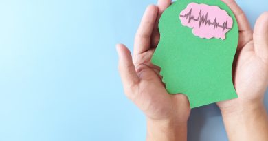 Two hands hold a green paper cutout shaped like a human head against a light blue background, with a pink brain shape inside the head displaying a heartbeat-style waveform line.