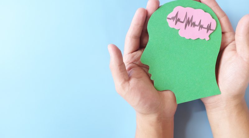 Two hands hold a green paper cutout shaped like a human head against a light blue background, with a pink brain shape inside the head displaying a heartbeat-style waveform line.