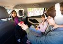 Mother offers apple to son in car while daughter watches tablet with headphones, capturing a modern family road trip balancing healthy snacks and connected entertainment