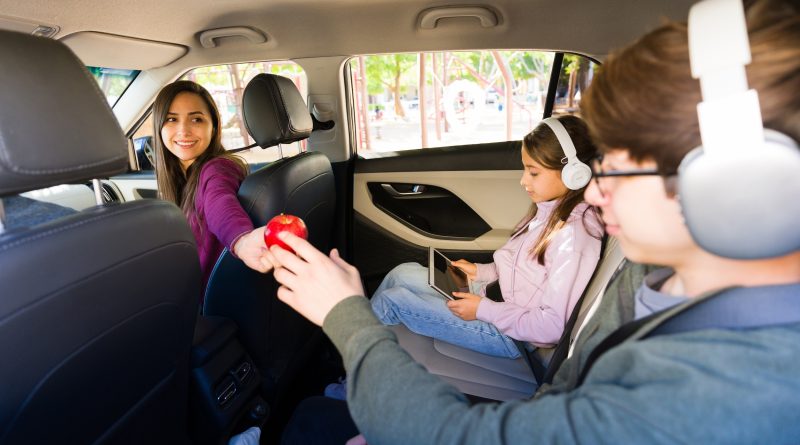 Mother offers apple to son in car while daughter watches tablet with headphones, capturing a modern family road trip balancing healthy snacks and connected entertainment