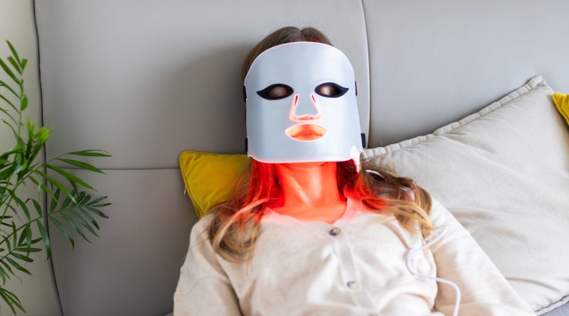 Woman using LED light therapy mask at home. Relaxing on couch.