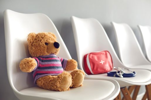 First-aid kit, stethoscope and cute teddy bear on white chairs in waiting room of children's medical center or pediatric clinic. Concept of kids' visit to hospital to see pediatrician or family doctor.