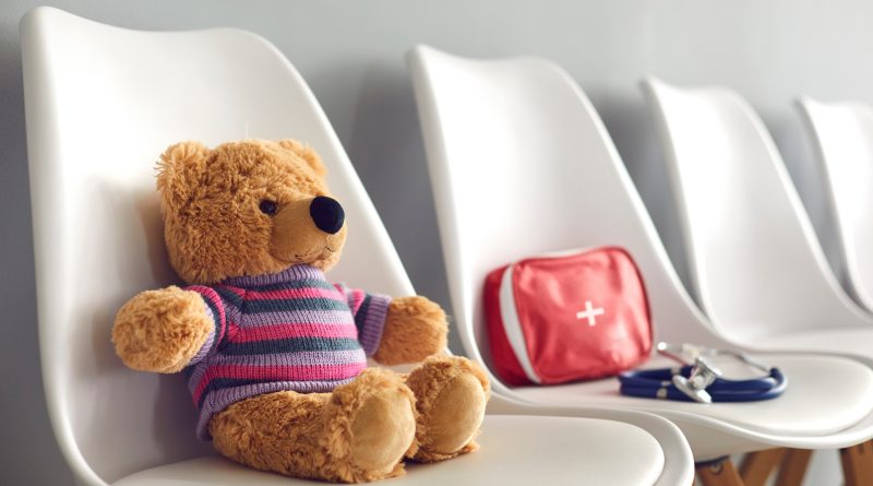 First-aid kit, stethoscope and cute teddy bear on white chairs in waiting room of children's medical center or pediatric clinic. Concept of kids' visit to hospital to see pediatrician or family doctor.