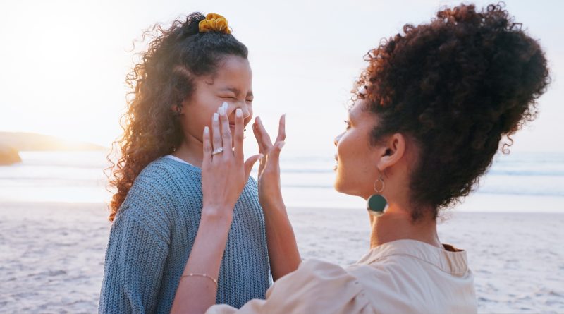 Mother, girl and sunscreen application at beach for uv protection, care and love on vacation in summer. Mom, child and lotion for face, skincare and bonding with smile by ocean on holiday.