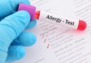 Close-up of a gloved hand holding a blood sample tube labeled “Allergy-Test” above a medical allergy testing form. The form includes checked boxes for IgE level testing and allergy screening for food and inhalant allergens, illustrating allergy diagnosis and laboratory testing.